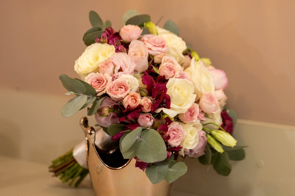 A close-up of a floral bouquet arranged with a mix of pale pink roses, white roses, deep burgundy flowers, and sprigs of eucalyptus leaves, positioned in a silver vase on a beige surface against a sof