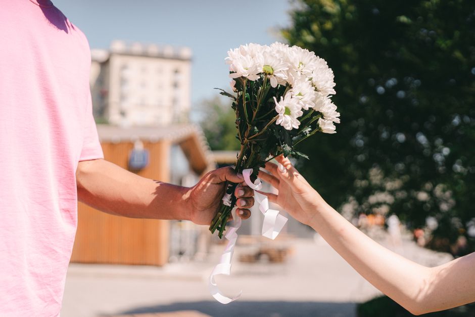 A person in a pink shirt hands over a bouquet of fresh white chrysanthemums with green stems and leaves to another individual outdoors. The bouquet is wrapped with white ribbons, and the arrangement i
