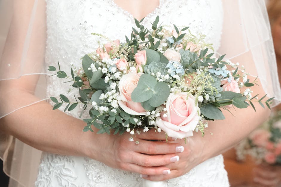 A bride holding a bouquet arrangement featuring soft pink roses, white baby's breath, green eucalyptus leaves, and succulent accents, wrapped in white tissue paper. The bouquet is positioned in front 