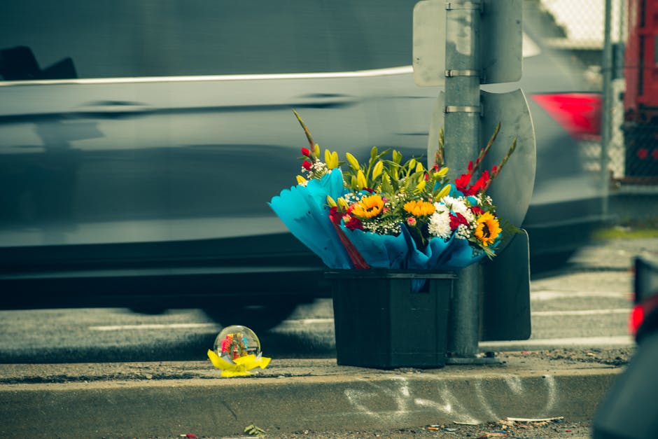 A bouquet of fresh flowers placed on a pavement next to a traffic light post, featuring a mix of yellow sunflowers, red and white blossoms, and green leafy foliage, wrapped in bright blue paper with a