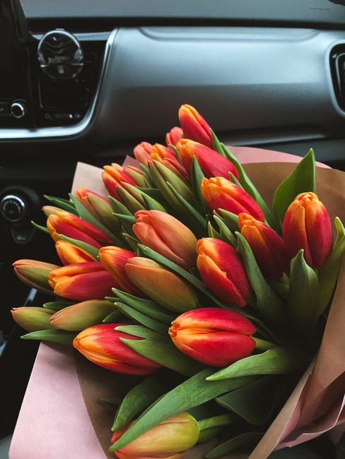 A vibrant bouquet of fresh tulips with bright orange and red petals, complemented by green stems and leaves, wrapped in light pink and brown paper. The bouquet is being held inside a car, positioned i